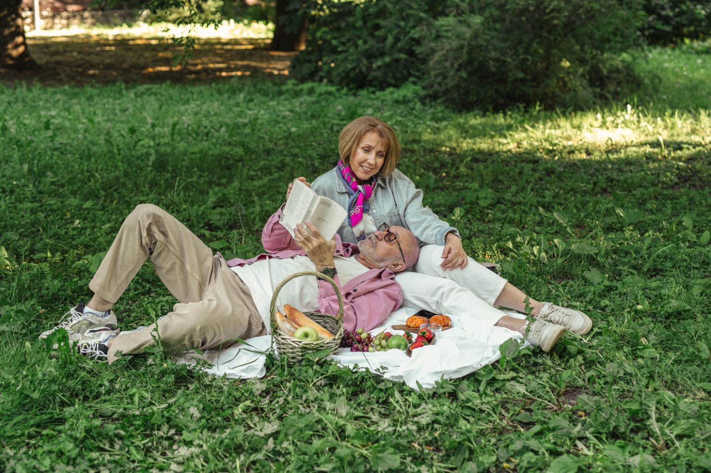 Retired couple having a picnic in a park: he lies reading a book, his head on her lap, she smiles, sitting behind him